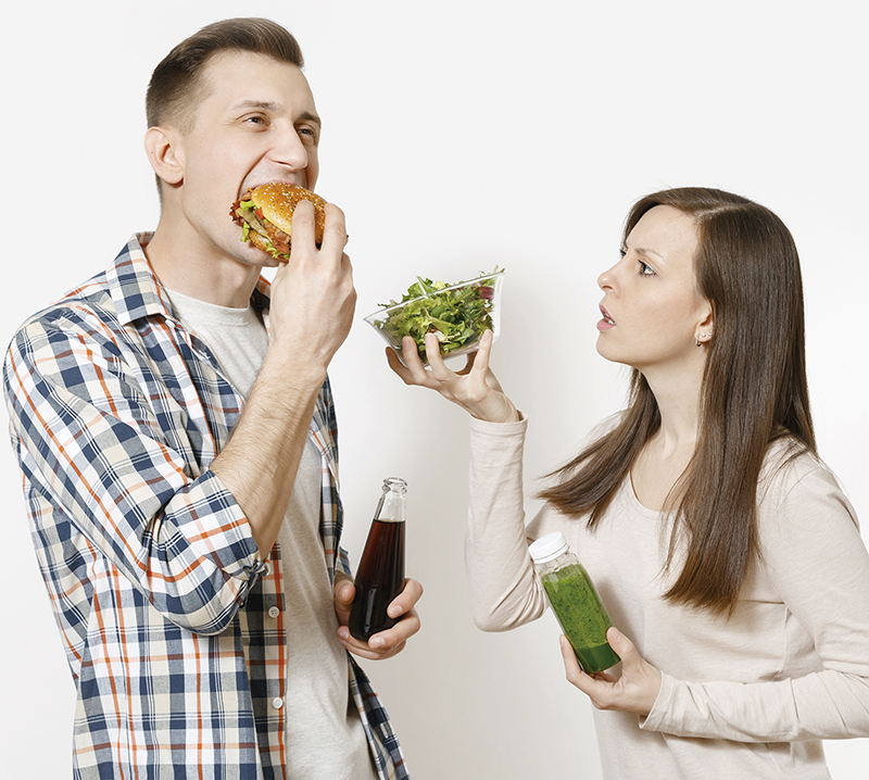 Couple man and woman standing with green detox smoothies, salad in glass bowl, burger, cola in glass bottle isolated on white background. Proper nutrition, healthy lifestyle, fast food, choice concept